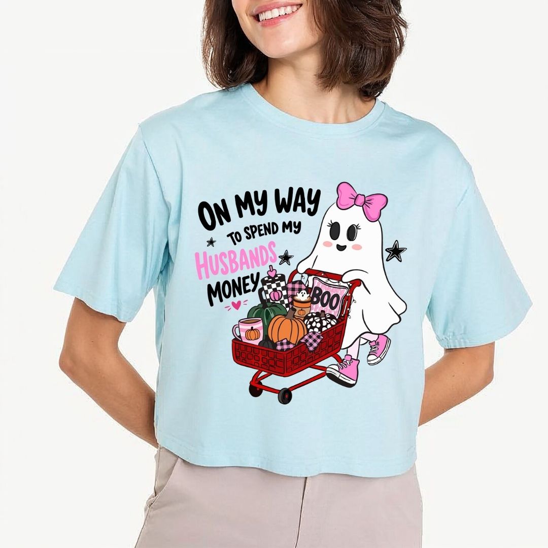 A person wearing a light blue crewneck t-shirt printed with the "On My Way to Spend My Husbands Money" ghost Halloween design.