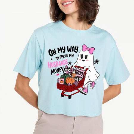 A person wearing a light blue crewneck t-shirt printed with the "On My Way to Spend My Husbands Money" ghost Halloween design.