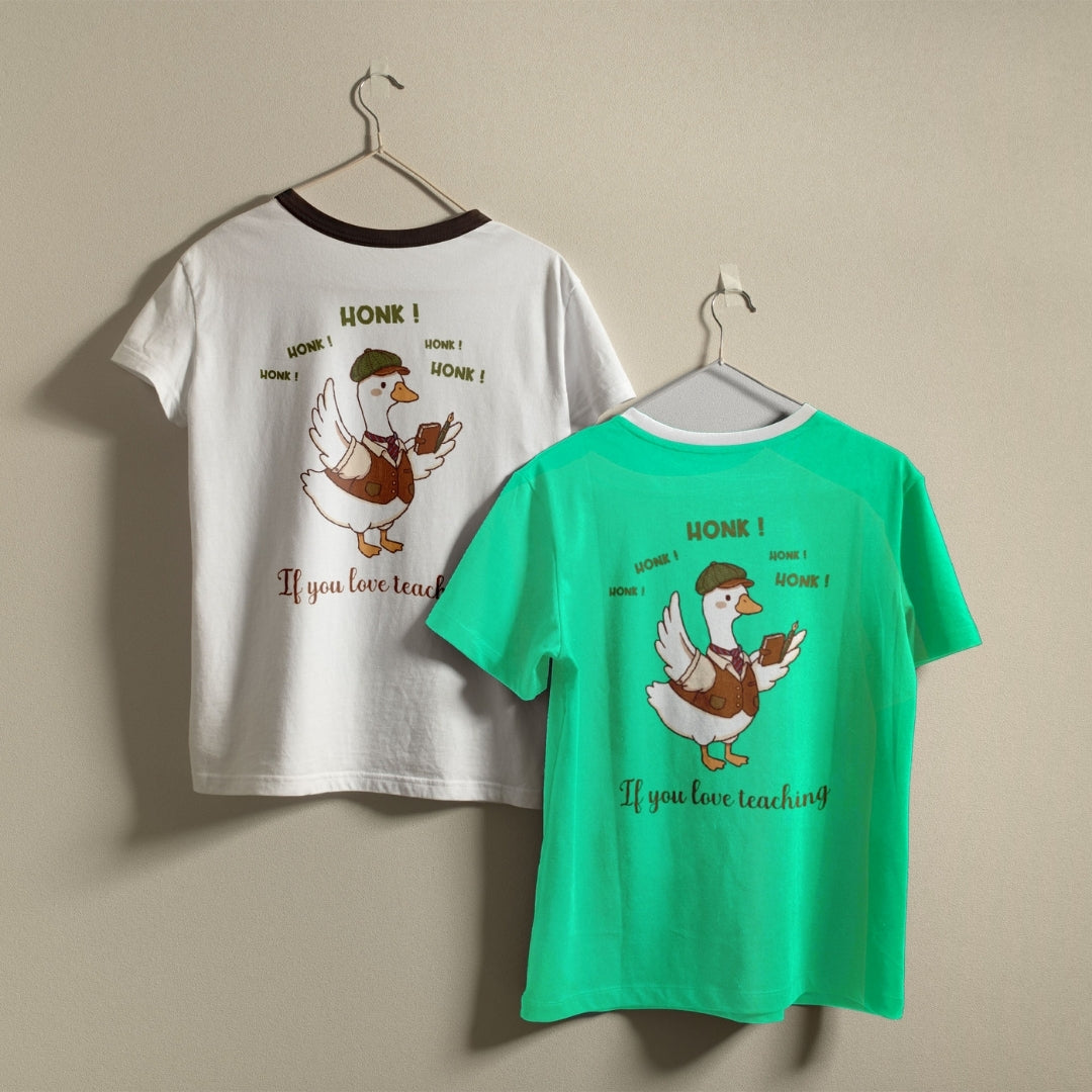 White and bright green t-shirts hanging on a wall, both displaying the humorous teacher duck graphic printed on the back.