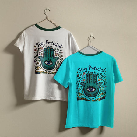 White and turquoise t-shirts hanging on a wall, both displaying the colorful Hamsa hand "Stay Protected" graphic printed on the back.