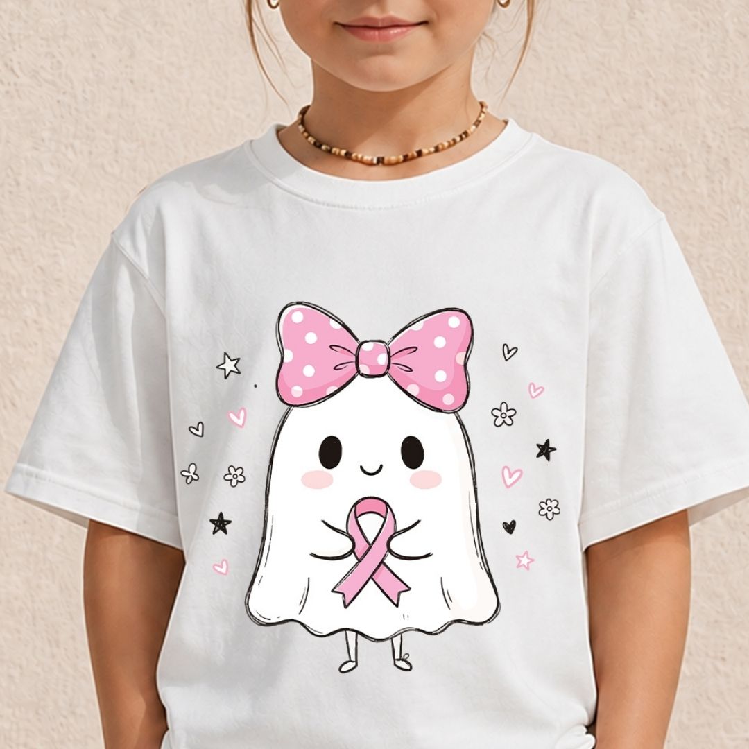 A child wearing a white short-sleeve T-shirt with a cute cartoon ghost graphic holding a pink ribbon, featuring a pink polka-dot bow and small pink and black decorative accents. The child is shown from the neck down against a light beige textured wall.