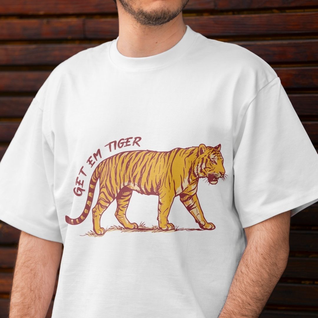 A man wearing a white short-sleeve t-shirt with a graphic of a walking tiger and the text 'GET 'EM TIGER' in red and orange, set against a dark wooden background.