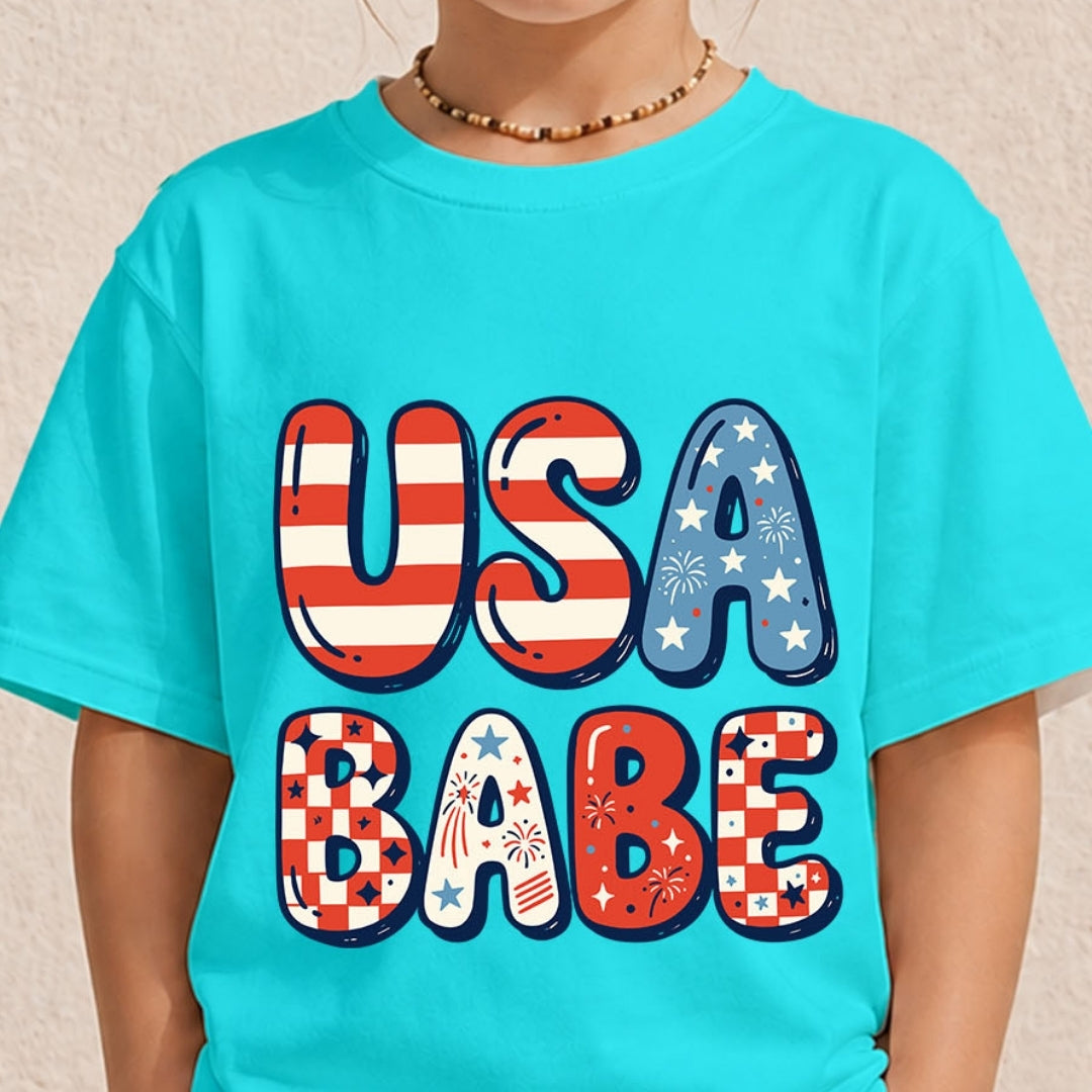 A close-up of a person wearing a bright turquoise short-sleeve T-shirt with large decorative text reading 'USA BABE' in red, white, and blue patriotic patterns. The photo is cropped from the lower face to the torso against a light neutral wall.
