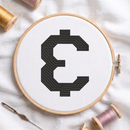 A wooden embroidery hoop holding white fabric with a large black embroidered letter 'E' in a pixelated, retro block pattern. The hoop is styled on a white fabric background with vintage spools of thread and a metal needle.