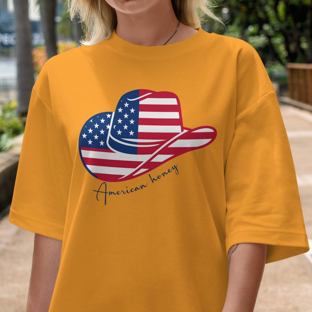 Person wearing a mustard yellow t-shirt with a graphic of a cowboy hat and American flag, text 'American honey'.