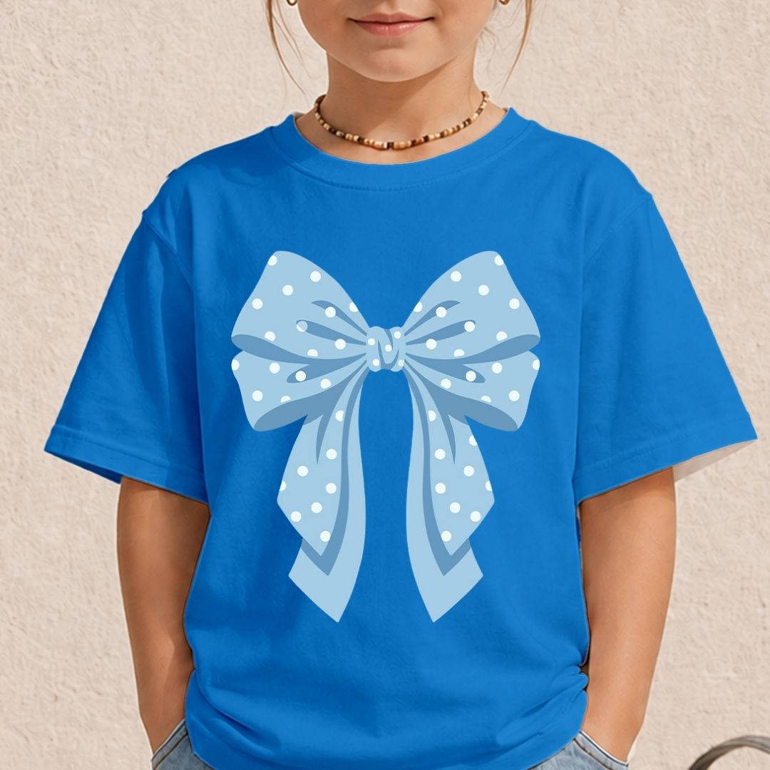 A child wearing a bright blue short-sleeve t-shirt with a large light blue and white polka dot bow graphic printed on the front, set against a textured beige wall.