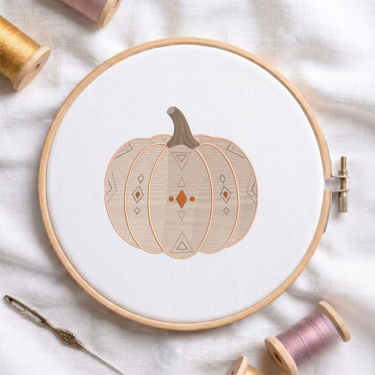 Geometric patterned pumpkin machine embroidery design displayed in a wooden hoop on white fabric with thread spools and a needle