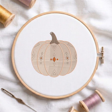 Geometric patterned pumpkin machine embroidery design displayed in a wooden hoop on white fabric with thread spools and a needle