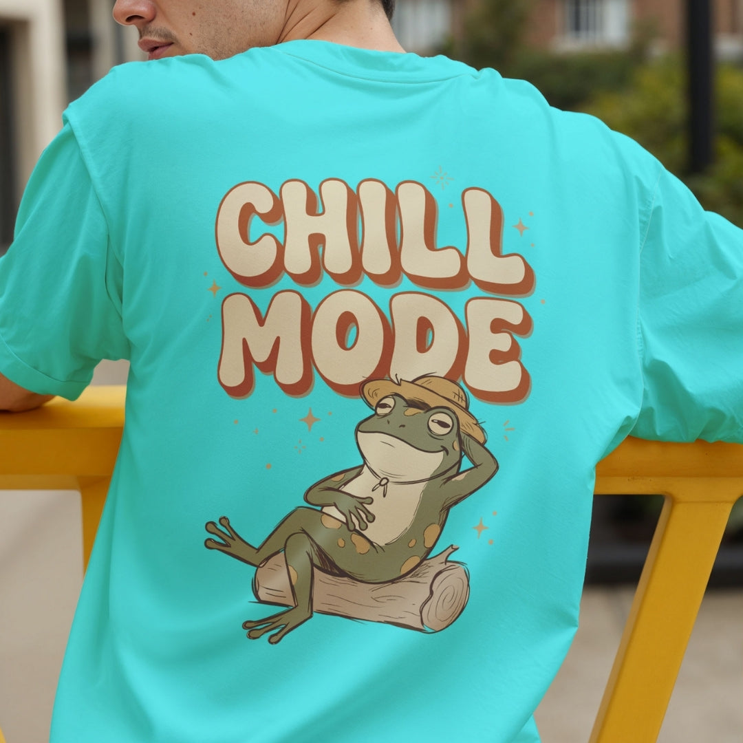 A person wearing a bright turquoise T-shirt shown from the back, featuring a large graphic of a relaxed frog sitting on a log with the words “CHILL MODE” in bold cream letters with brown outlines.
