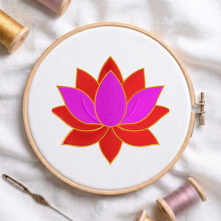 A circular embroidery hoop holding white fabric with a stylized lotus flower design in bright pink and red outlined with gold stitching. Spools of thread, a needle, and soft fabric are arranged around it in a styled flat-lay.