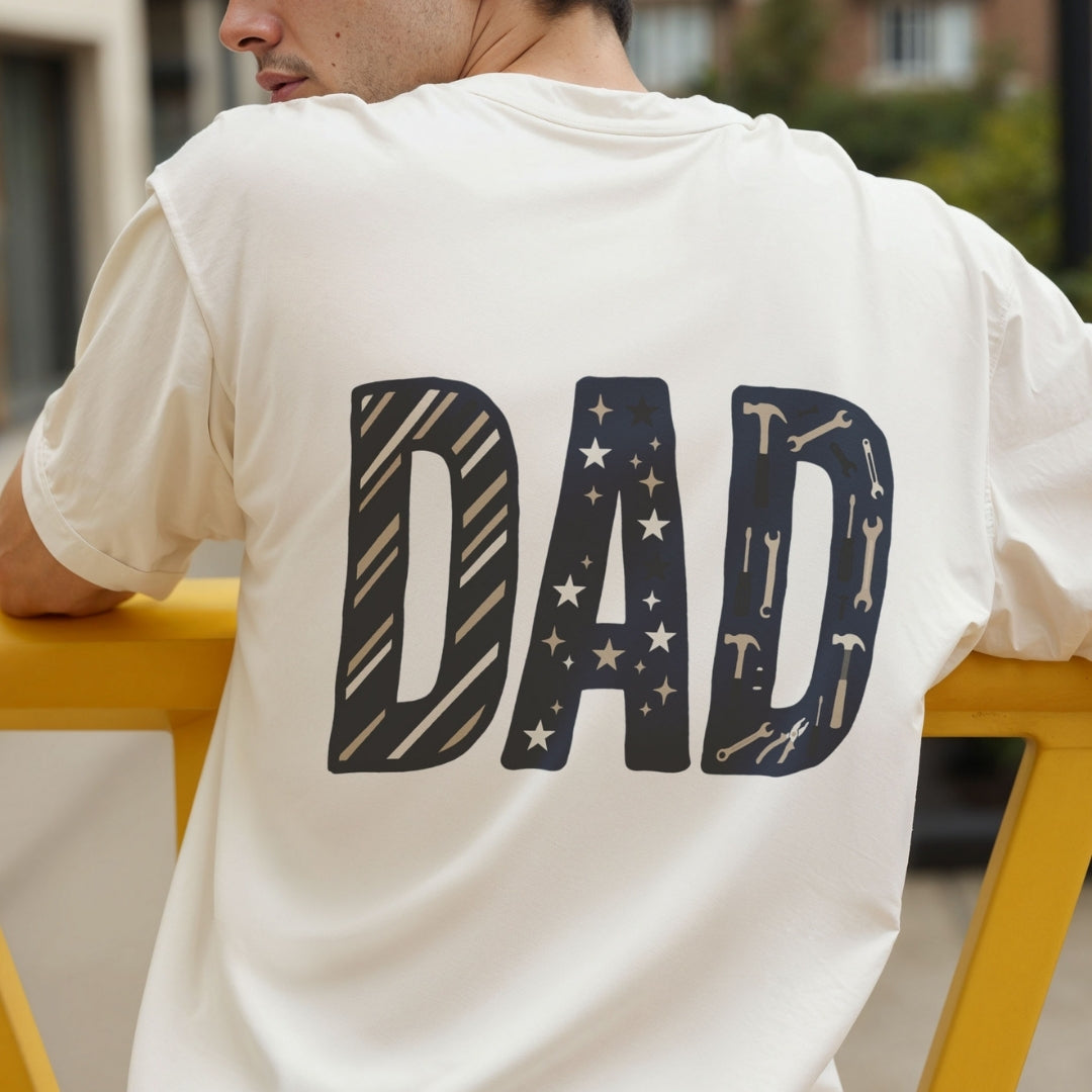 A man wearing a cream-colored t-shirt from the back, featuring the word 'DAD' in large navy blue letters decorated with stars and tool patterns.