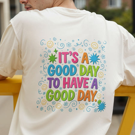 A man wearing a white t-shirt from behind, showing a colorful graphic print on the back that reads 'IT'S A GOOD DAY TO HAVE A GOOD DAY.' surrounded by smiley faces, stars, and swirls.