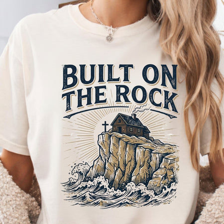 White t-shirt with 'Built on the Rock' graphic design featuring a house on a cliff.