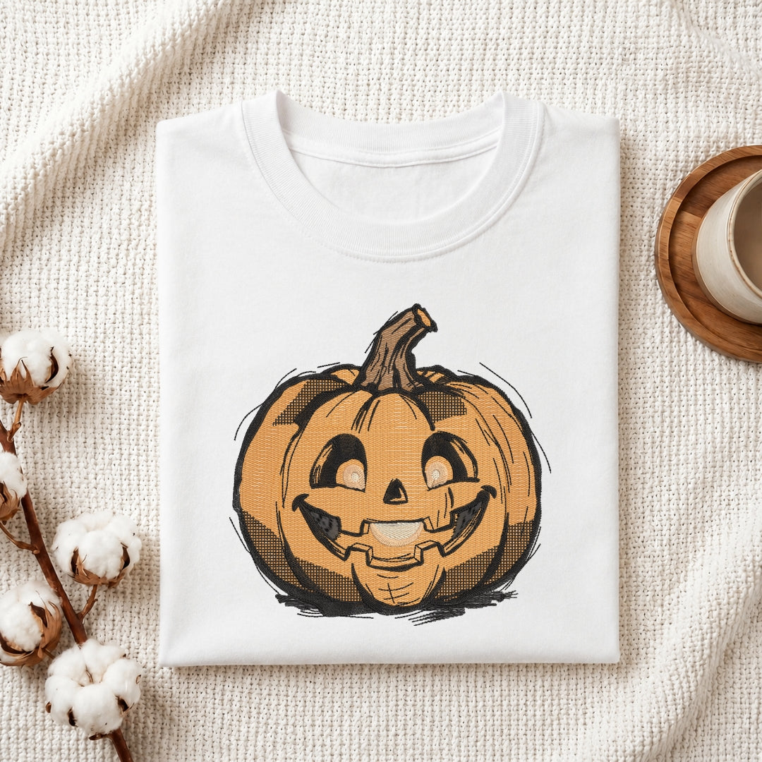 A folded white crewneck t-shirt featuring a graphic of a smiling jack-o'-lantern pumpkin, styled on a textured cream fabric with cotton bolls and a wooden cup in the background.