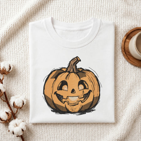 A folded white crewneck t-shirt featuring a graphic of a smiling jack-o'-lantern pumpkin, styled on a textured cream fabric with cotton bolls and a wooden cup in the background.