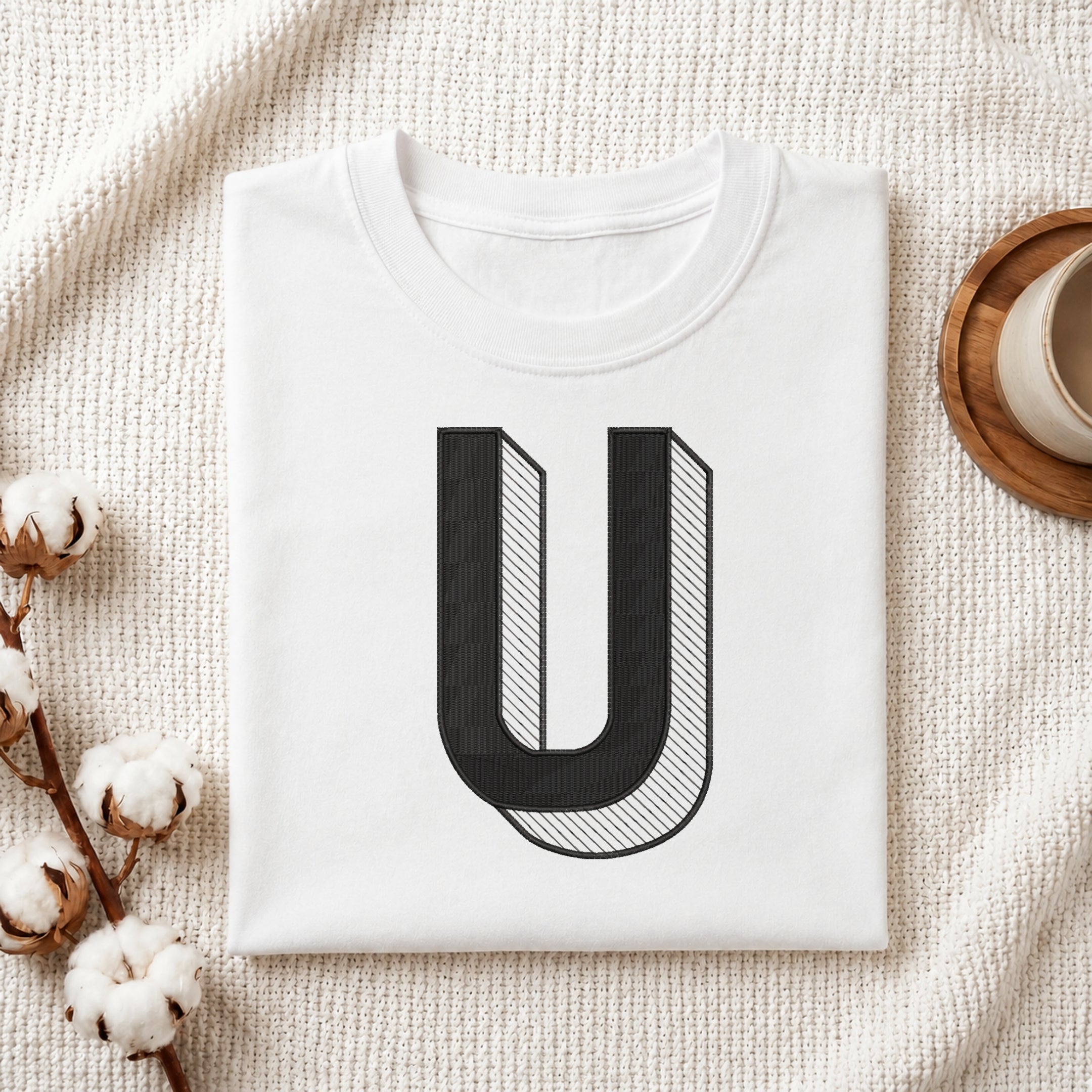 A neatly folded white crewneck t-shirt featuring a stitched 3D block letter "U" design, styled with cotton branches and a wooden coaster.