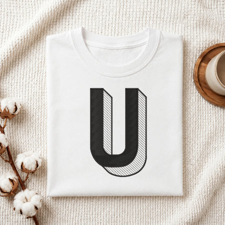 A neatly folded white crewneck t-shirt featuring a stitched 3D block letter "U" design, styled with cotton branches and a wooden coaster.