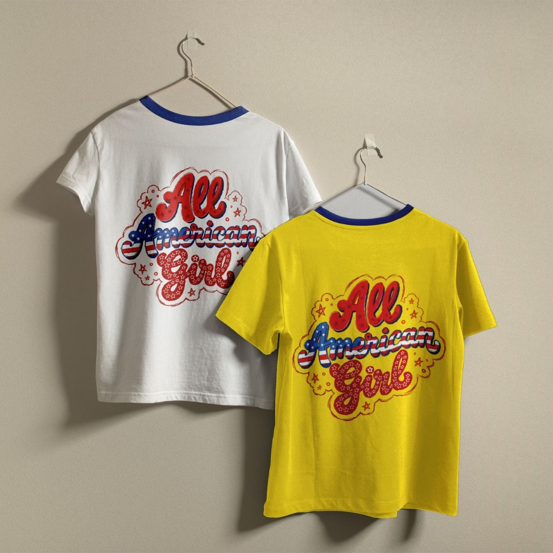 Two vintage-style ringer t-shirts with 'All American Girl' graphics hanging on hangers against a plain beige wall. One shirt is white and the other is bright yellow, both featuring blue ribbed neck trims and a colorful retro graphic on the back.