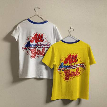 Two vintage-style ringer t-shirts with 'All American Girl' graphics hanging on hangers against a plain beige wall. One shirt is white and the other is bright yellow, both featuring blue ribbed neck trims and a colorful retro graphic on the back.