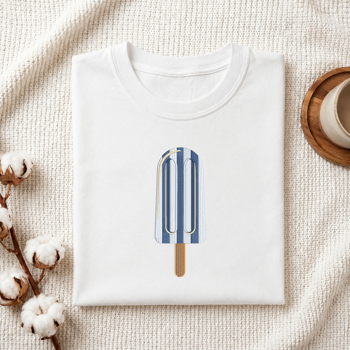 Folded white cotton t-shirt mockup featuring a blue and white striped ice cream popsicle machine embroidery design, styled with cotton stems and a wooden cup.