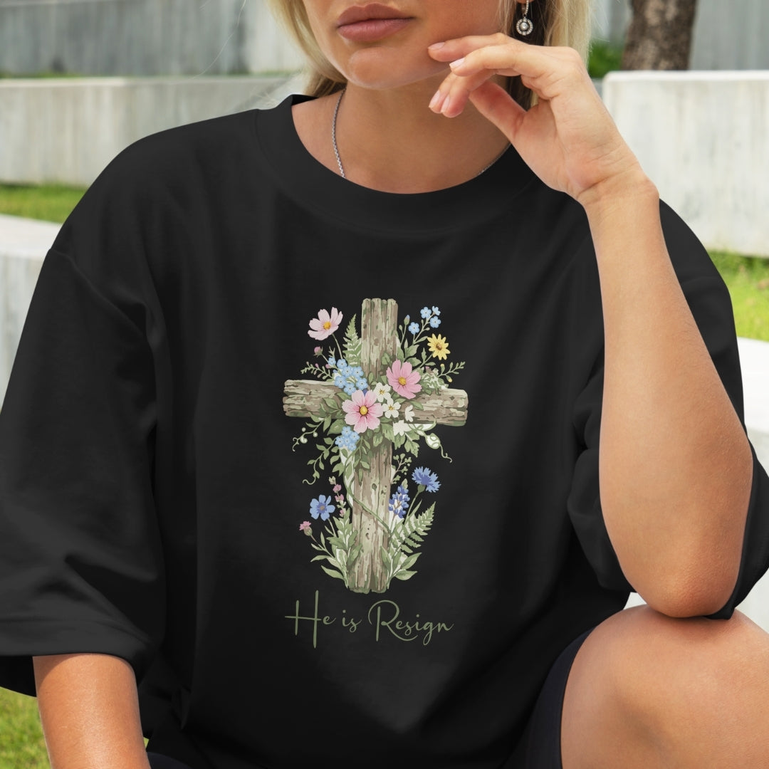 Person wearing a black t-shirt with a floral cross design and 'He is Risen' text.
