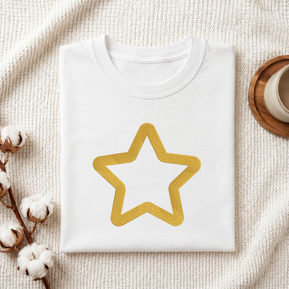 Folded white cotton t-shirt mockup featuring a golden star outline machine embroidery design, styled with cotton stems and a wooden cup