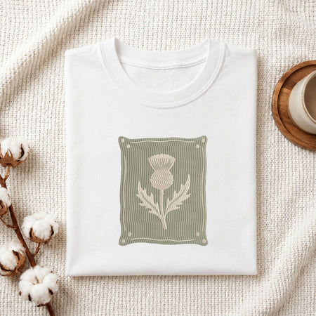A folded white crewneck t-shirt with a central graphic of a beige thistle on a muted sage green striped background, styled on a textured cream blanket with cotton bolls and a wooden cup.