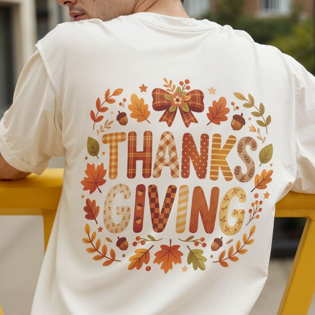 A person viewed from behind wearing a white T-shirt with a large autumn-themed 'THANKSGIVING' graphic on the back, featuring fall leaves, acorns, stars, and patterned lettering, posed on a yellow bench.