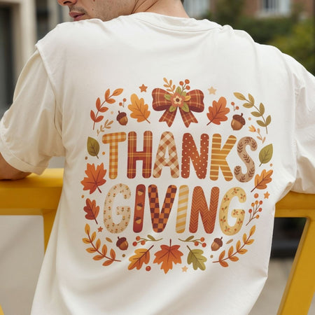 A person viewed from behind wearing a white T-shirt with a large autumn-themed 'THANKSGIVING' graphic on the back, featuring fall leaves, acorns, stars, and patterned lettering, posed on a yellow bench.