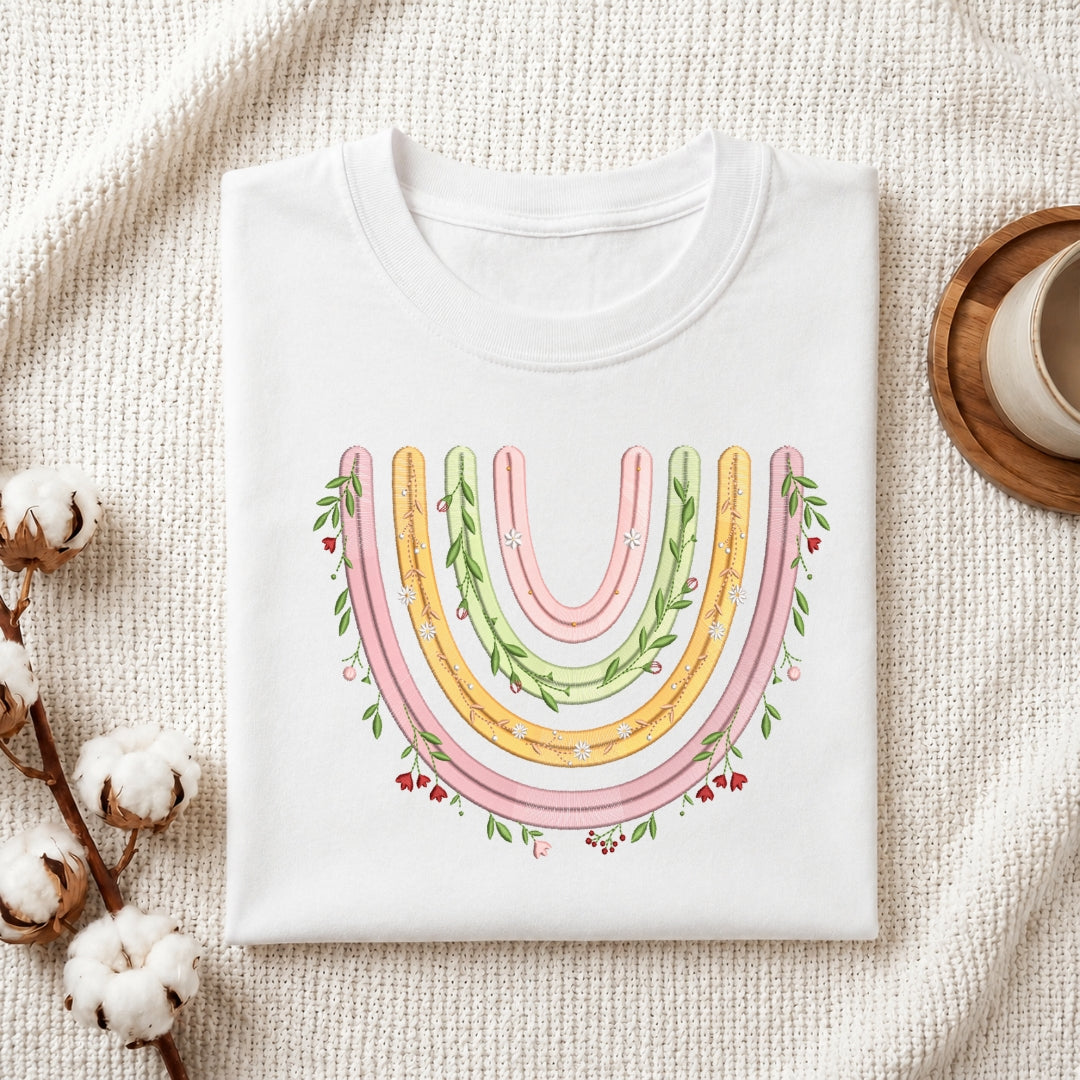 A folded white crewneck t-shirt with a colorful watercolor-style rainbow graphic featuring green leaves and small red berries, styled on a textured cream fabric with cotton bolls and a wooden cup.
