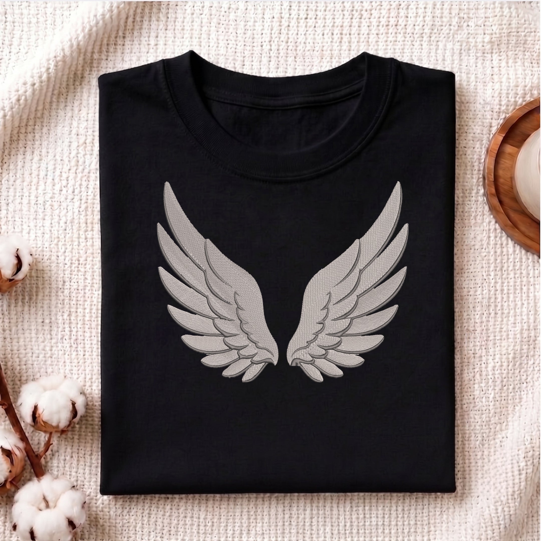 A folded black crewneck t-shirt with a large gray embroidered angel wings graphic on the front, displayed on a textured beige blanket with cotton bolls and a wooden tray.