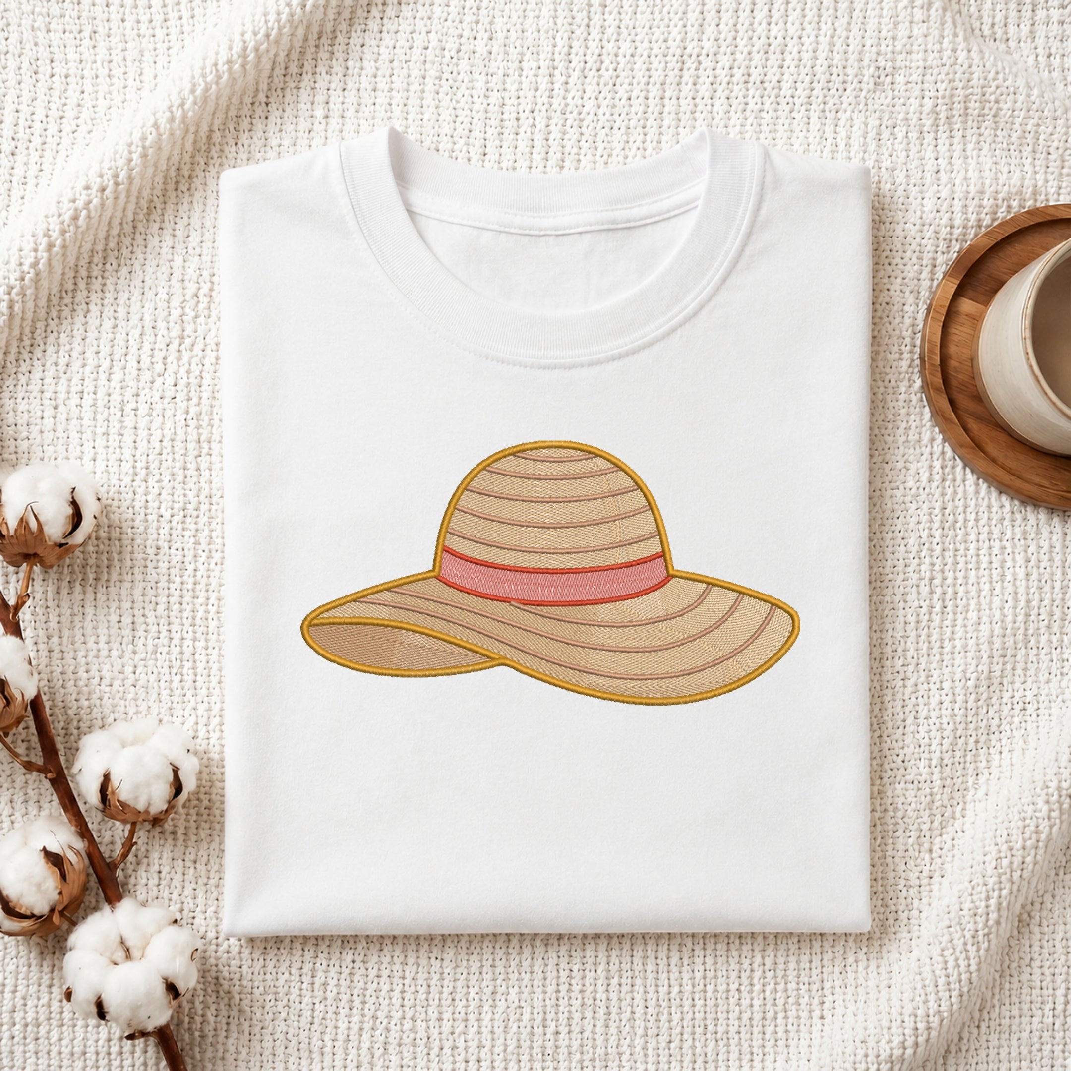 A folded white T-shirt laid flat on a textured cream fabric background, featuring an embroidered straw hat design with a pink band. Cotton bolls and part of a wooden cup are styled around the shirt as props.
