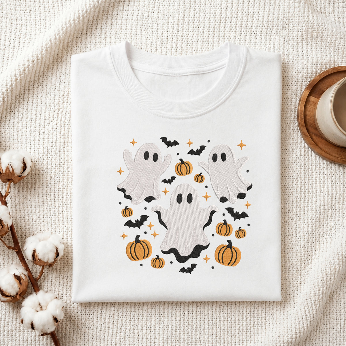 A folded white crewneck t-shirt with a Halloween graphic print featuring three ghosts, pumpkins, bats, and stars, styled on a textured cream fabric with cotton stems and a wooden cup.