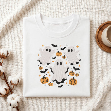 A folded white crewneck t-shirt with a Halloween graphic print featuring three ghosts, pumpkins, bats, and stars, styled on a textured cream fabric with cotton stems and a wooden cup.
