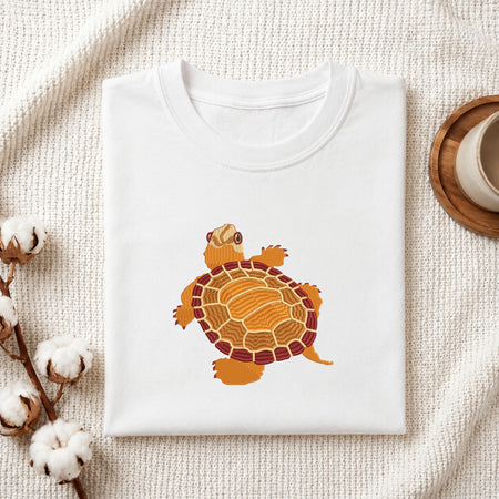Folded white cotton t-shirt mockup featuring a turtle machine embroidery design, styled with cotton stems and a wooden cup
