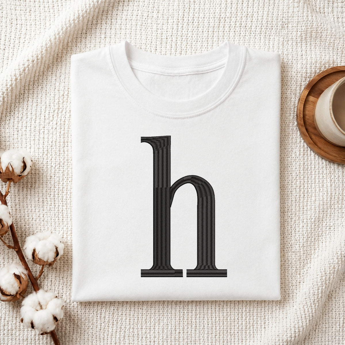 A folded white t-shirt with a large black serif letter 'h' printed on the front, styled on a textured cream fabric background with cotton bolls and a wooden cup.