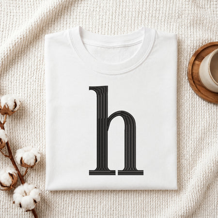A folded white t-shirt with a large black serif letter 'h' printed on the front, styled on a textured cream fabric background with cotton bolls and a wooden cup.