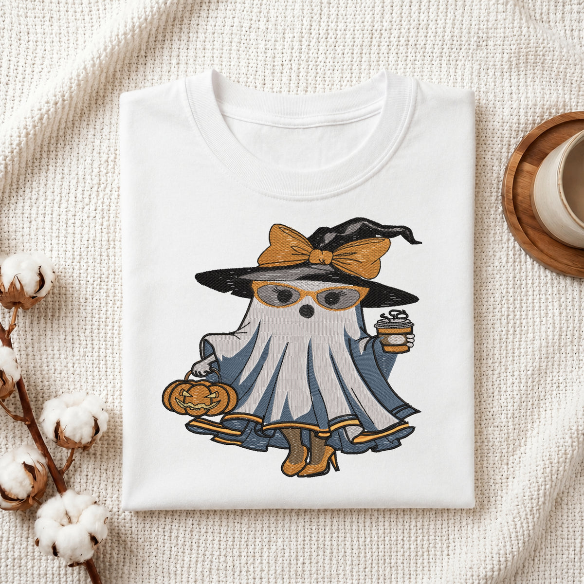 Folded white t-shirt mockup with an embroidered fashionable ghost wearing a witch hat, glasses, and high heels while holding coffee and a pumpkin.