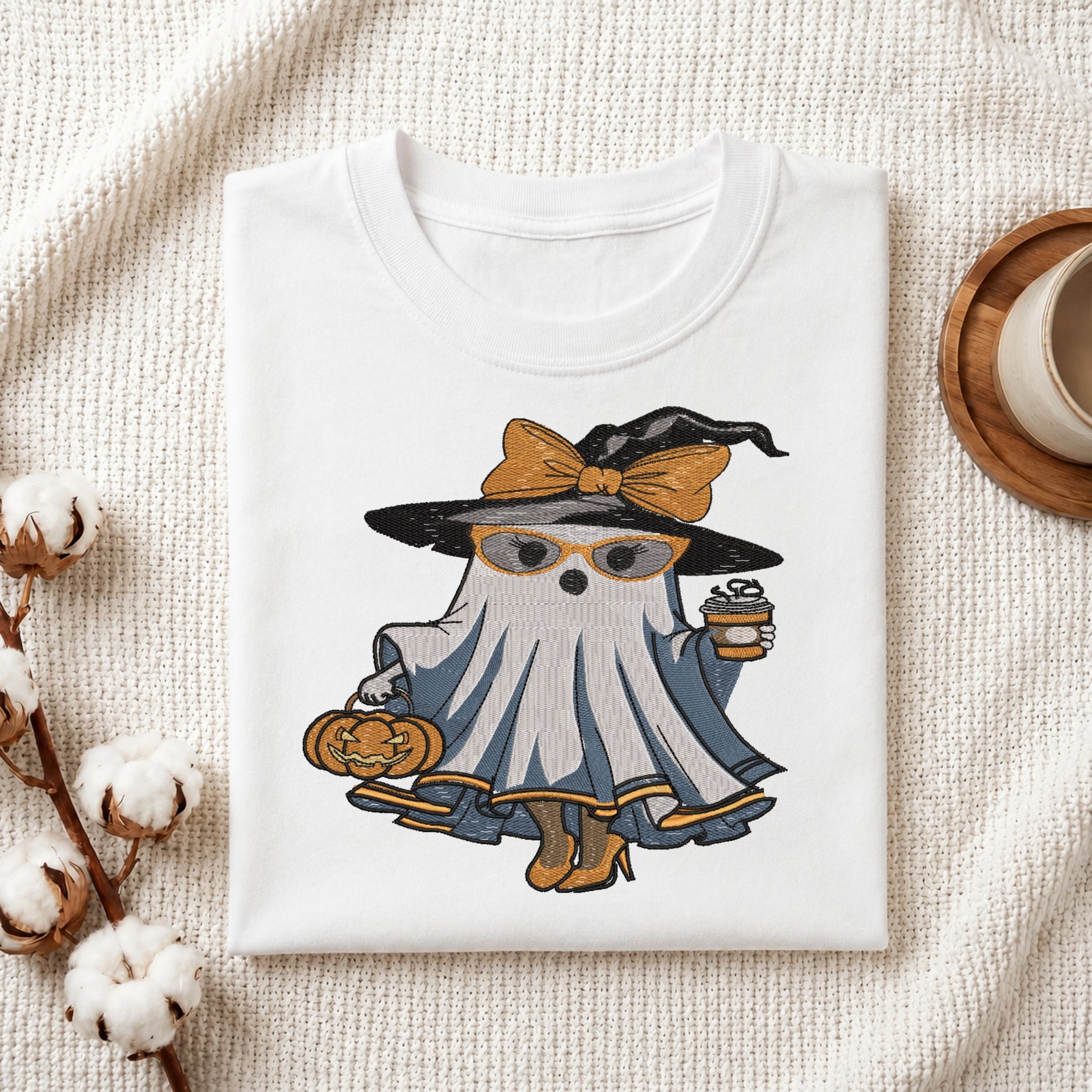 Folded white t-shirt mockup with an embroidered fashionable ghost wearing a witch hat, glasses, and high heels while holding coffee and a pumpkin.