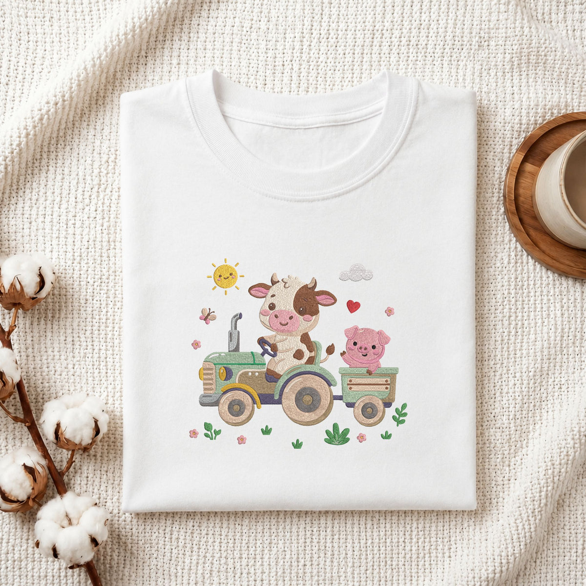 Folded white cotton t-shirt mockup featuring a cute cow and pig machine embroidery design, styled with cotton stems and a wooden cup   