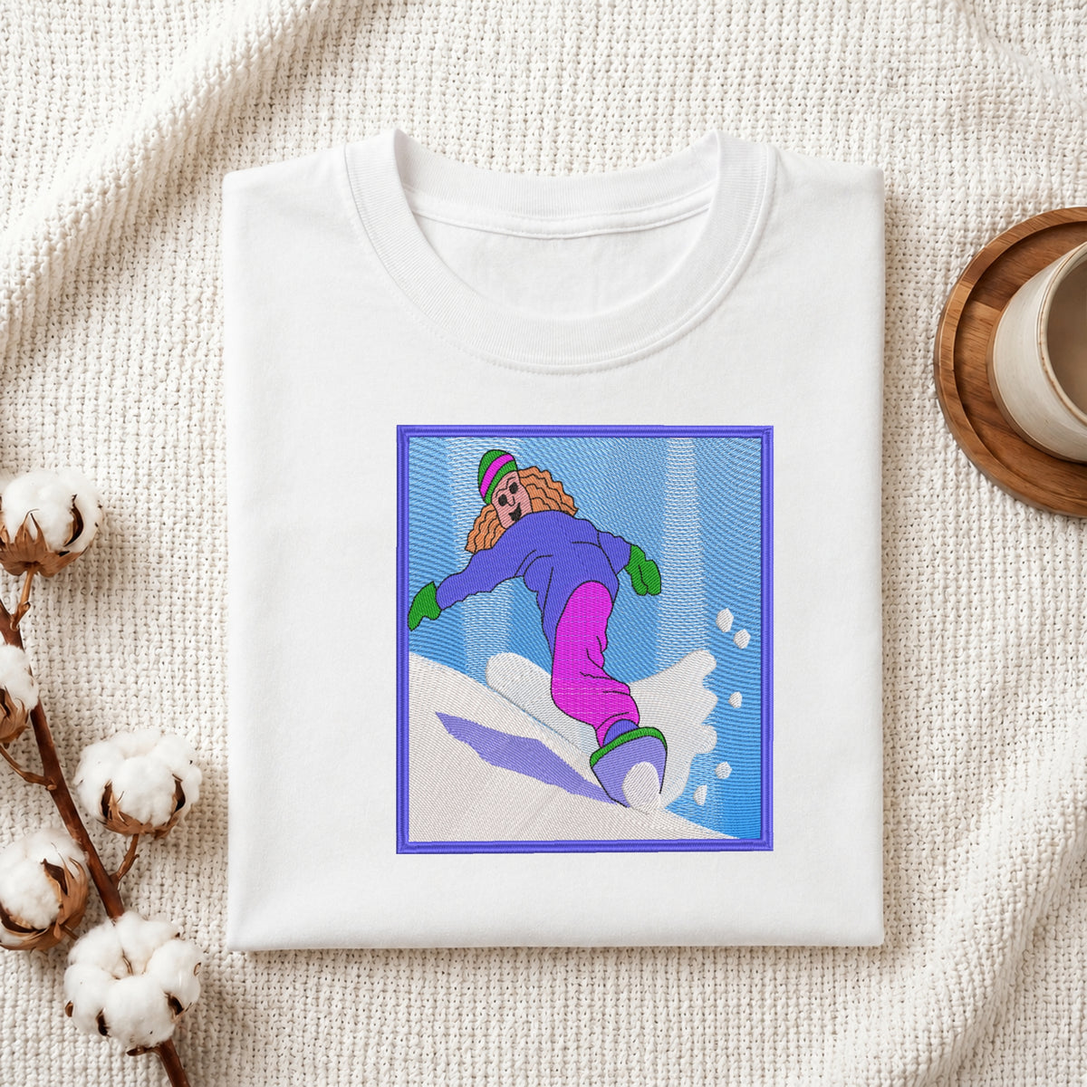 A folded white crewneck t-shirt with a colorful graphic print of a woman snowboarding, styled on a textured cream knit blanket with cotton bolls and a coffee cup.