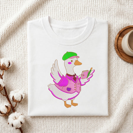 A white t-shirt featuring an embroidered design of a duck wearing a green cap, a pink vest, and holding a small notebook and pen.