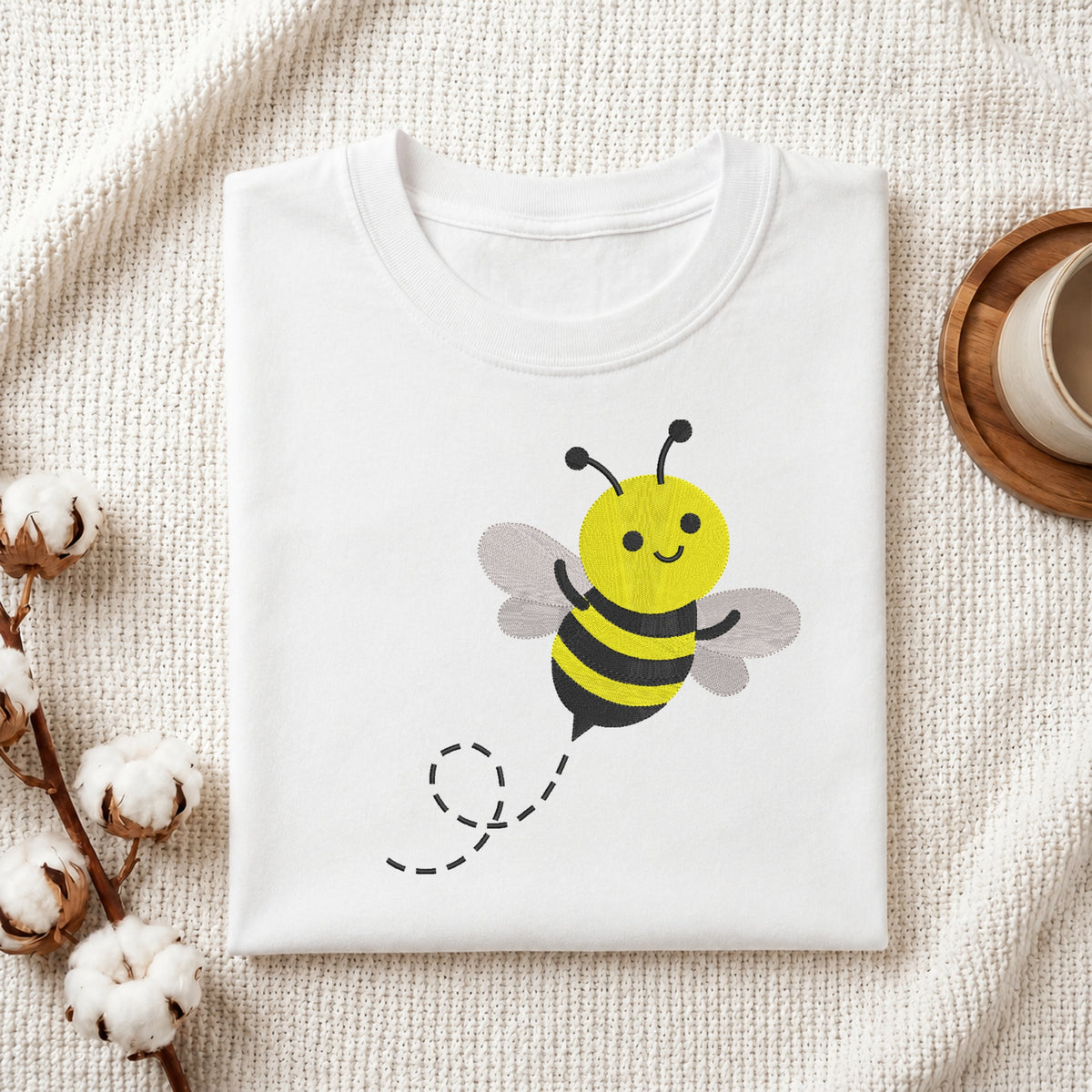 A folded white t-shirt with a cute cartoon bee graphic, styled on a textured cream fabric with cotton bolls and a wooden cup in the background.