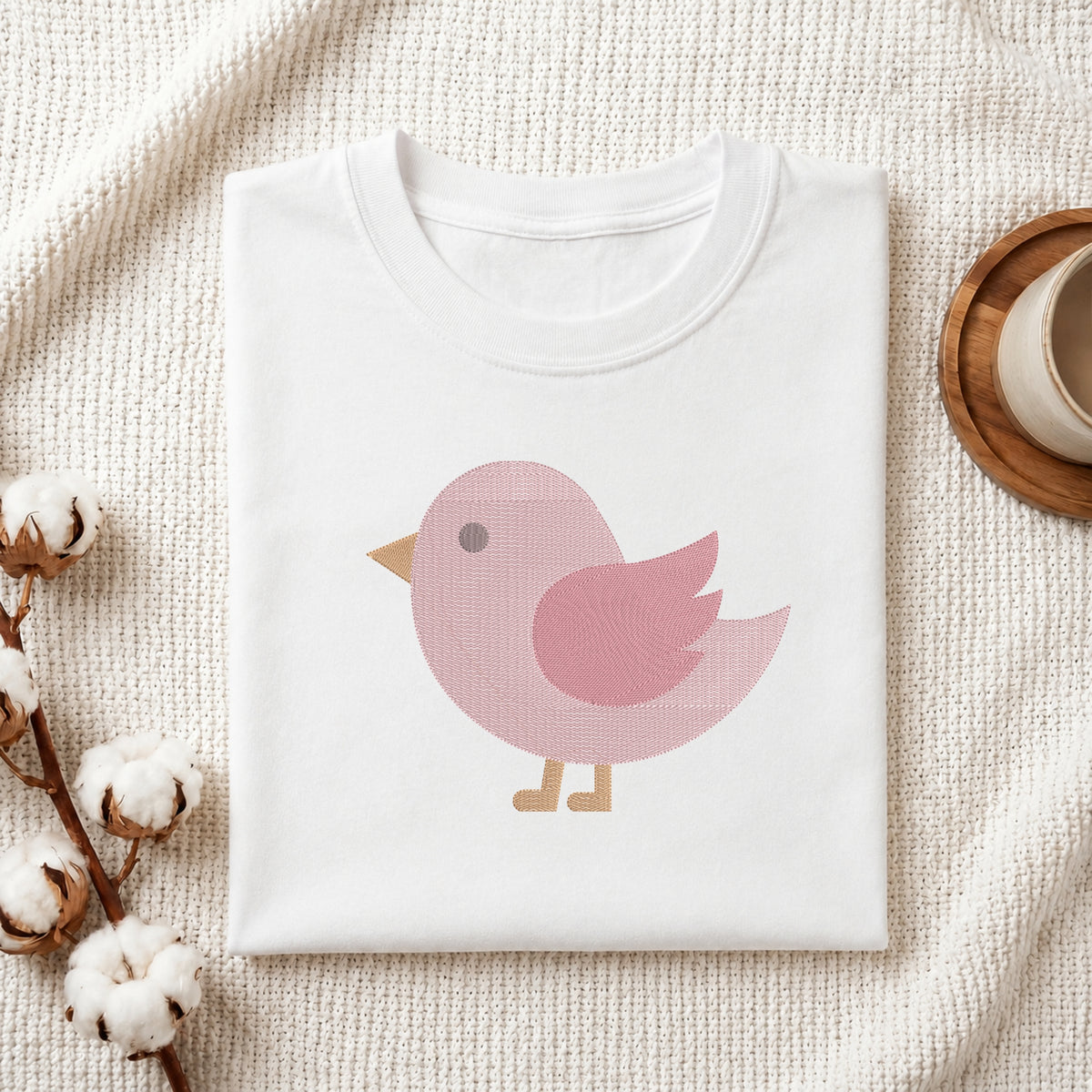 A folded white crewneck t-shirt with a pink bird graphic, styled on a textured cream knit blanket with cotton bolls and a wooden cup in the background.