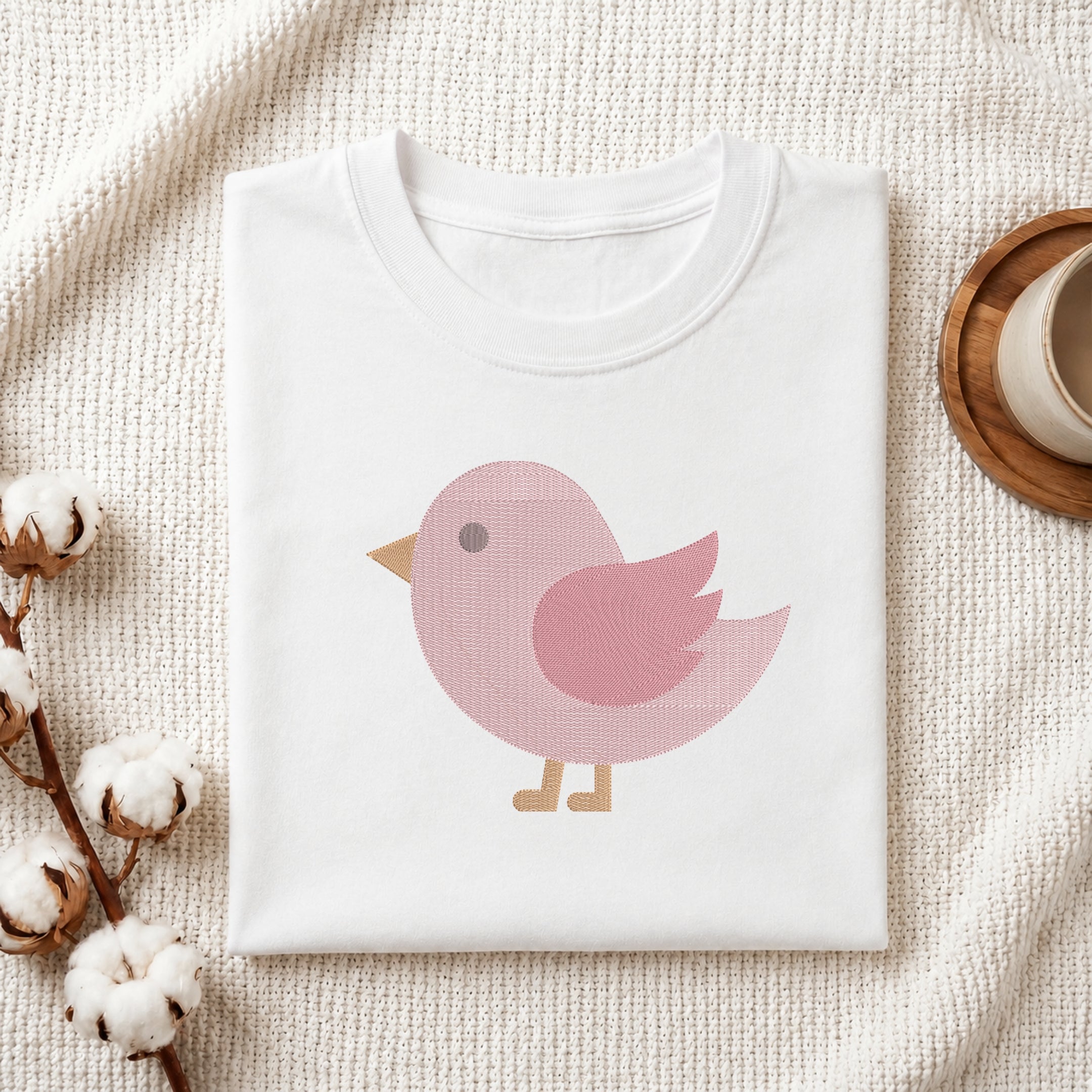 A folded white crewneck t-shirt with a pink bird graphic, styled on a textured cream knit blanket with cotton bolls and a wooden cup in the background.