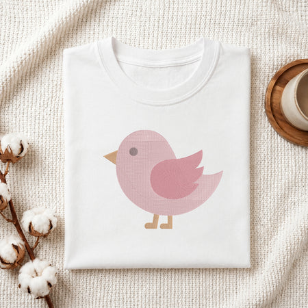 A folded white crewneck t-shirt with a pink bird graphic, styled on a textured cream knit blanket with cotton bolls and a wooden cup in the background.