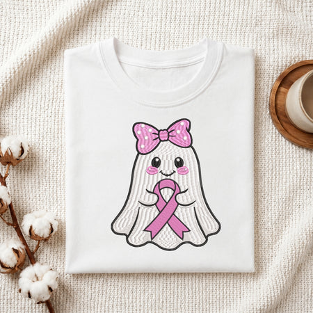 A folded white t-shirt with a cute cartoon ghost graphic holding a pink awareness ribbon, styled on a textured cream fabric with cotton bolls and a wooden cup in the background.
