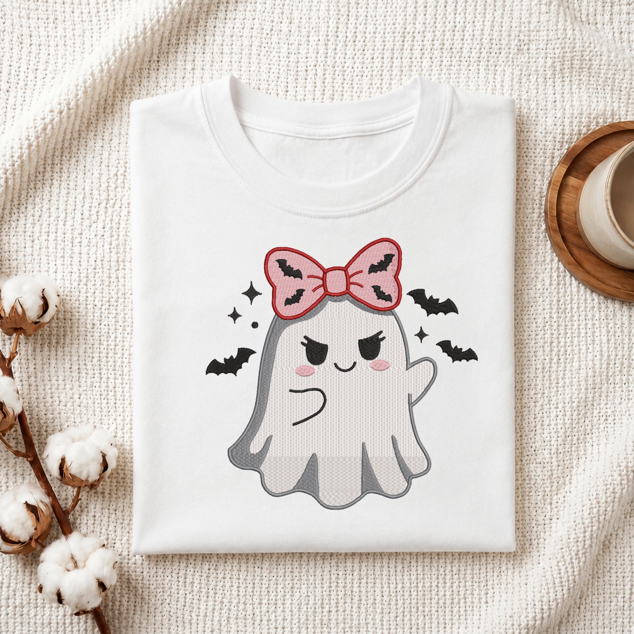 A neatly folded white t-shirt featuring the embroidered spooky ghost with bat bow design, displayed on a textured white knit background with cotton stems and a wooden cup.
