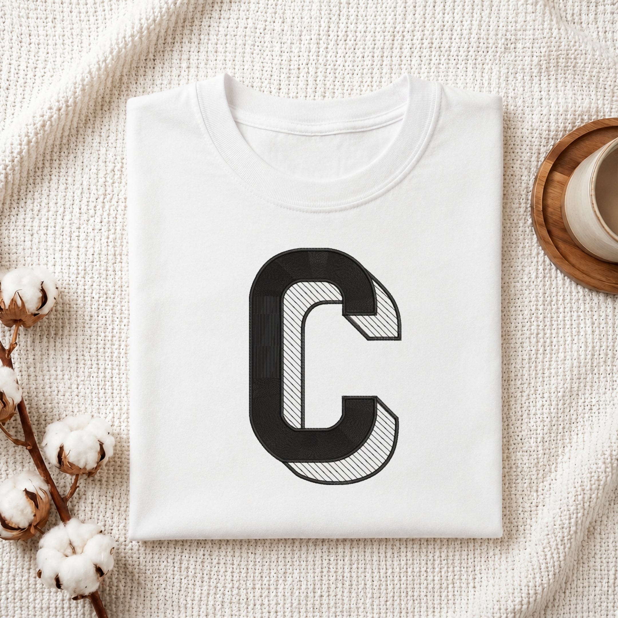 A folded white crewneck t-shirt with a large black graphic letter 'C' on the front, styled on a textured cream knit blanket with cotton stems and a wooden cup in the background.