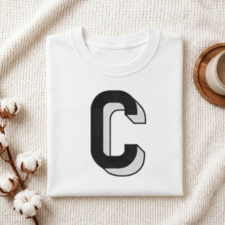 A folded white crewneck t-shirt with a large black graphic letter 'C' on the front, styled on a textured cream knit blanket with cotton stems and a wooden cup in the background.